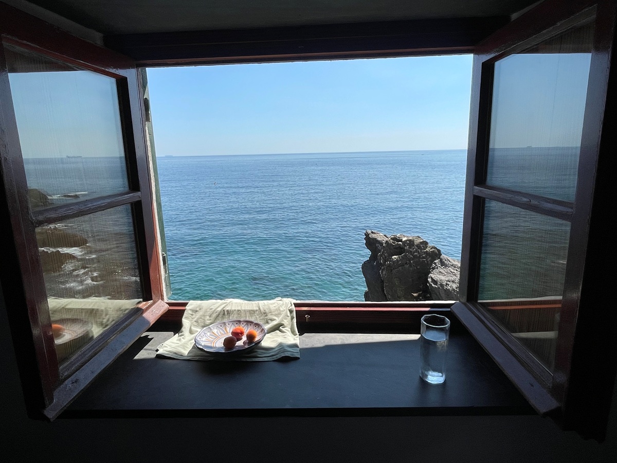 An open wooden window in the Baracca in Nervi, Italy, looking out over the Mediterranean Sea. A ceramic dish of fruit and a glass of water sit on the sill, and a weathered rock rises from the water beyond.